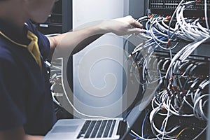 Young man connecting wires in server cabinet while working with laptop in data center