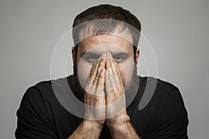 The young man covered his face with his hands. Brunette in a black T-shirt. Disturbing emotions. Gray background