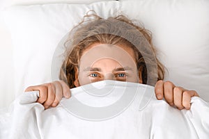 Young man covering his face with blanket while lying on pillow. Bedtime