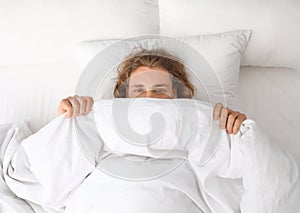 Young man covering his face with blanket while lying on pillow, top view
