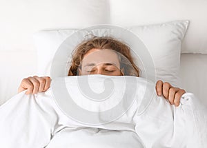 Young man covering his face with blanket while sleeping on pillow. Bedtime