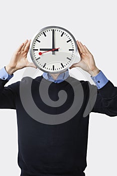 Young man covering his face with clock standing against white background