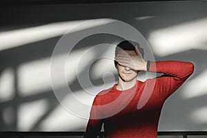 Young man covering his face with his hand in a red sweater on a white background with shadows from the light. Copy, empty space