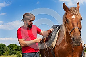 Young man in cowboy hat saddling his brown horse