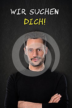 Young man with crossed arms and `Wir suchen dich` text on a blackboard background. Translation: `We are looking for you`