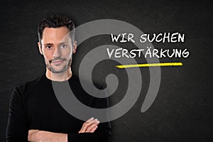 Young man with crossed arms and `Wir suchen dich` text on a blackboard background. Translation: `We are looking for you`