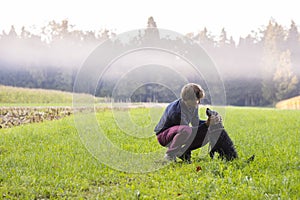 Young man crouching to pet his black dog in a beautiful green me