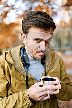 Young man with a cup of coffee in his hands looks frowningly while standing in the park