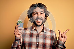 Young man with curly hair and beard holding small cactus plant pot over yellow background very happy pointing with hand and finger