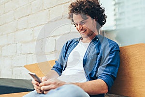 Young man with curly hair stitting outdoors typing on mobile phone. Happy male using smart phone application for searching