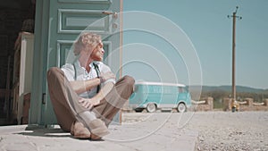 Young man with curly hair in white shirt and brown trousers sitting by the door on a dusty road. Action. Hot summer