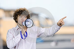 Young man with curly hair in white shirt speaks in megaphone pointing with index finger.