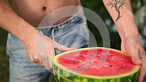 Young man cuts knife into slices, big ripe red watermelon, on street, Sunny day