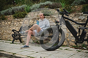 young man cyclist in sportswear, drinking water from sports bottle in the city, sitting on a bench, relaxing after