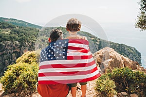 Young man dad father and his son school boy kid standing on a rock cliff with US flag on shoulders and looking at sea