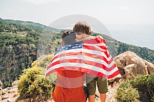 Young man dad father and his son school boy kid standing on a rock cliff with US flag on shoulders and looking at sea