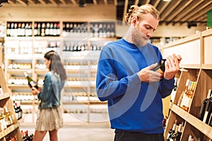 Young man in dark blue sweater thoughtfully reading label of wine in supermarket