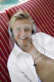 Young man on deckchair by pool listening to music on headphones portrait