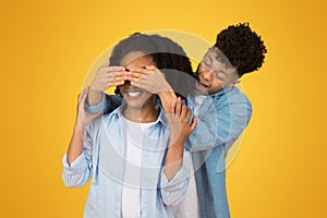 A young man in a denim shirt playfully covers a smiling woman's eyes from behind