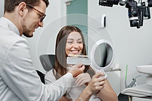 Young man dentist in white coat show veneers enamel color palette for beautiful patient woman