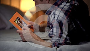 Young man in despair lying on bed looking at photo with girlfriend, sadness