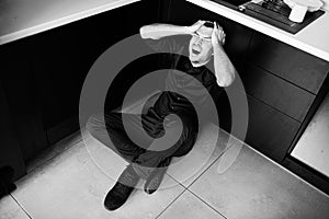 Young man in despair sit on kitchen`s floor and suffer. Hold hands on head. Scream and yell out looud. Mad businessman