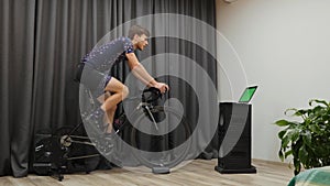 Young man doing cardio training on stationary bike trainer indoor. Professional cyclist preparing for competition and looking at g