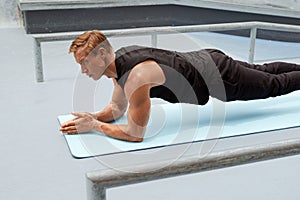 Young Man Doing Plank Exercise On Yoga Mat Against Concrete Wall Outdoors. Handsome Caucasian Sportsman.