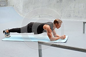 Young Man Doing Plank Exercise On Yoga Mat Against Concrete Wall Outdoors. Handsome Caucasian Sportsman.