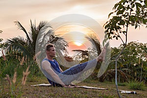 Young man doing yoga near the palm tree at sunset.