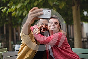 Young man with Down syndrome and his mentoring friend sitting and taking selfie outdoors in cafe