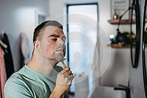 Young man with down syndrome learning how to shave, holding razor in hand, looking at mirror.