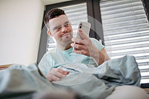 Young man with down syndrome lying in bed, looking at smartphone in morning. Morning routine for man with disability.