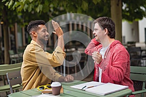 Young man with Down syndrome with mentoring friend sitting outdoors in cafe celebrating success.