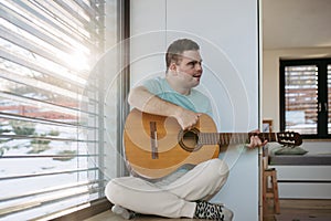 Young man with down syndrome playing acoustic guitar, sitting by window, holding and strumming guitar, making music.