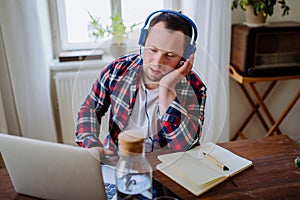 Young man with Down syndrome sitting at desk in office and using laptop, listening to music from headphones.