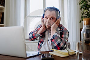 Young man with Down syndrome sitting at desk in office and using laptop, listening to music from headphones.