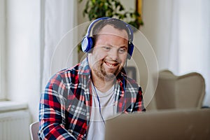 Young man with Down syndrome sitting at desk in office and using laptop, listening to music from headphones.