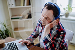 Young man with Down syndrome sitting at desk in office and using laptop, listening to music from headphones.