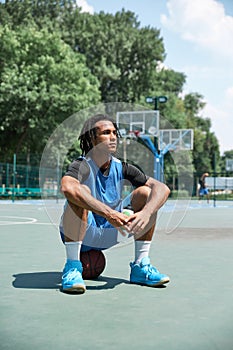 Young man with dreads, athlete in blue uniform sitting on basketball ball, resting after outdoors training on