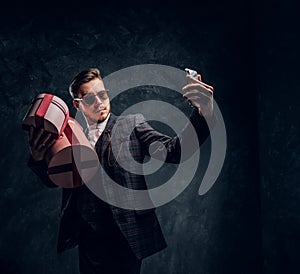A young man dressed in an elegant suit holding gifts and taking selfie by smartphone in a dark studio