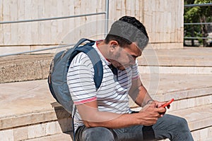 A young man dressed informally is sitting on a ladder, checking his phone photo