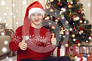 Young man dressed in retro sweater and sanat hat sit near christmas tree. New year eve, ornated fir tree. Winter holiday and love