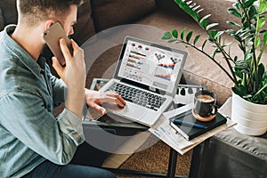 Young man, dressed in shirt, sits at home on couch at coffee table,uses laptop with graphs, charts, diagrams on screen