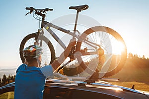 Young Man dresses modern cycling clothes and protective helmet installing his mountain bike on the car roof with sunset backlight