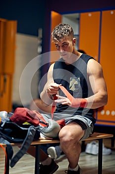 Young man in dressing room, sitting on bench, tying red tape around his hand, getting ready for workout