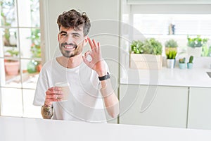 Young man drinking a coffee in a paper cup in the morning doing ok sign with fingers, excellent symbol