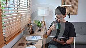 Young man drinking coffee and reading new on digital tablet at home.