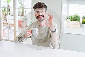 Young man drinking a cup of coffee in the morning doing ok sign with fingers, excellent symbol