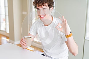 Young man drinking take away cup of coffee at home doing ok sign with fingers, excellent symbol